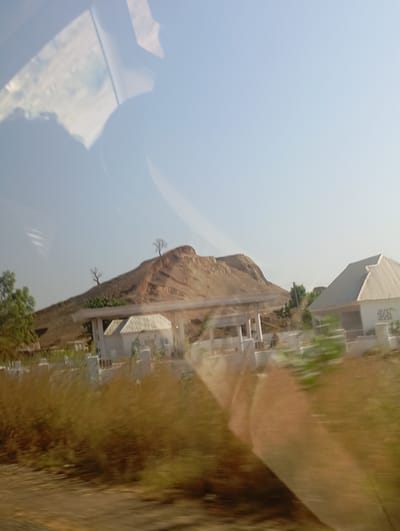 This daytime photograph, likely taken from a moving vehicle in Farei, Nigeria, captures a semi-rural landscape under a clear, light blue sky. A prominent, reddish-brown rocky hill, sparsely dotted with bare trees, rises in the background. At the base of this hill, in the midground, two light-colored buildings with white walls and sloped, possibly corrugated, roofs are visible. The structure on the left includes a covered, open-sided area supported by white pillars. White perimeter fencing or low walls surround the immediate vicinity of these buildings. The foreground is dominated by heavily motion-blurred, dry, yellowish-brown grass and scrub, with some green foliage present on the far left. Significant reflections, consistent with a vehicle's interior or ambient light, are present across the upper and central-right areas of the image. No individuals are discernible within the scene.