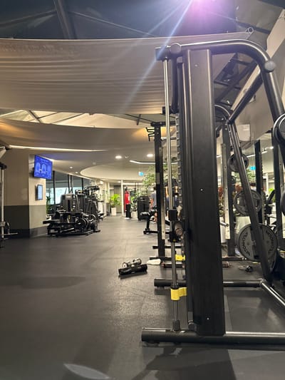 This image depicts the interior of a gym facility located in Bondi Beach, Australia. The scene is brightly illuminated by artificial overhead lighting, casting a lens flare in the upper right. The floor is covered in dark rubber matting.

In the immediate foreground on the right, a large, black metal multi-functional cable machine or power rack stands prominently, featuring weight plates, some branded "TECHNOGYM." Yellow safety latches are visible on its vertical support. On the floor directly in front of this machine, a black rolled yoga mat and two small dumbbells are present, along with a dark, wet-appearing stain or patch on the rubber flooring.

Moving into the midground and background, the gym extends into a spacious area filled with various black gym equipment, including weight machines, benches, and a red punching bag located towards the rear. Potted green plants are strategically placed throughout the facility. On the left wall, a blue-lit screen displays three distinct figures or icons, indicative of a workout interface or class schedule, with a narrower vertical screen below it. A male individual, with his back to the camera, is partially visible in the mid-right background near additional equipment. The facility features curved architectural elements, high ceilings with a light-colored fabric canopy structure, and large windows or glass partitions along the left side. Blue and white exit signs are visible in the distance. No active interactions or specific actions beyond the presumed use of the facility are discernible.