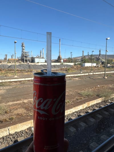 The scene depicts a clear daytime view from what appears to be a moving train or stationary train window, overlooking railway tracks towards an industrial complex in Sidi Kacem, Morocco. In the immediate foreground, a person's hand, partially visible, holds a chilled red Coca-Cola can with condensation droplets. A clear plastic straw is inserted vertically into the can's opened tab. The can displays "Coca-Cola" and "GOÛT ORIGINAL" in white script. The midground features multiple parallel railway tracks with concrete sleepers, bordered by a concrete platform or retaining wall and dry, sparse vegetation on gravel. Several dark grey streetlights or utility poles are positioned alongside the tracks, and multiple thin overhead wires stretch horizontally across the blue sky. The background is dominated by a sprawling industrial facility, identified as an oil refinery or petrochemical plant, characterized by numerous tall metal towers, interconnected piping, storage tanks, and smokestacks under a clear blue sky. Large grey aggregate piles are visible between the railway line and the industrial plant, with sporadic green vegetation and palm trees.