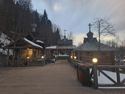 The image depicts a complex of wooden buildings connected by a wooden boardwalk in Vzglyadnevo, Russia. The buildings feature traditional Russian architectural elements, including steeply pitched roofs and crosses atop the structures. Snow is visible on the roofs and in the surrounding area, suggesting a winter setting. The boardwalk is lined with wooden railings and illuminated by spherical light fixtures. Three trash cans are positioned near the boardwalk, adding a mundane element to the otherwise historical scene. Bare trees and a hillside backdrop contribute to the natural environment. The sky is overcast, casting a muted light on the scene. People are visible in one of the buildings in the distance.