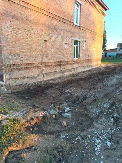 The image shows a construction site next to a brick building. The building is made of red bricks and has two windows, one on the upper floor and one on the lower floor. The lower part of the building has a concrete foundation, and there is exposed rebar and concrete where some of the foundation has been removed or is under repair.
The ground in front of the building is disturbed earth, with tire tracks visible, indicating recent activity with heavy machinery. There are piles of debris, including broken bricks and concrete, scattered around the site. Patches of grass and weeds are growing in some areas, suggesting the construction work may have been ongoing or recently started. The scene appears to be outdoors, with a clear blue sky visible in the background, indicating fair weather. The sunlight casts long shadows, suggesting it is either early morning or late afternoon.
In the background, other buildings and a fence are visible, along with some trees. The overall impression is one of ongoing construction or renovation work in a somewhat rural or suburban setting. There is no visible text in the image. The mood is utilitarian, focused on the construction activity.