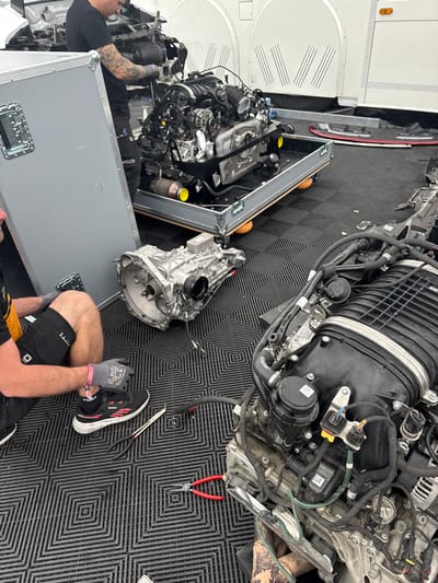 The scene depicts a mechanic working on a disassembled car engine, located on a black tiled floor. The engine is lying on its side with numerous wires and hoses visible. A second engine, partially disassembled, sits on a pallet nearby. A man in a black shirt and blue jeans is working on the engine, with his back turned towards the camera. There are a set of pliers, a pair of red pliers, and a few other tools scattered around the area. The location of the image is the Circuit de Spa-Francorchamps in Stavelot, Belgium, based on the provided coordinates. The mechanic's actions and the disassembled engines suggest that this is a racing or motorsports environment.