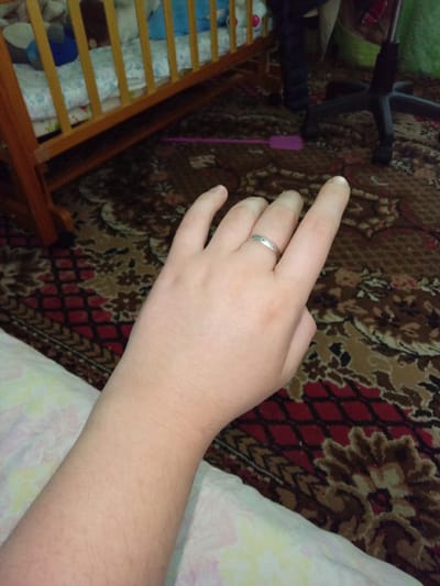 The image shows a close-up view of a person's arm and hand, resting on a patterned carpet. The person is wearing a silver ring on their left ring finger. In the background, a wooden crib with bedding and toys can be seen. A black office chair and a pink object on the floor are also visible. The scene appears to be indoors, with the lighting suggesting it is daytime. The location is identified as Kitskany, Moldova.