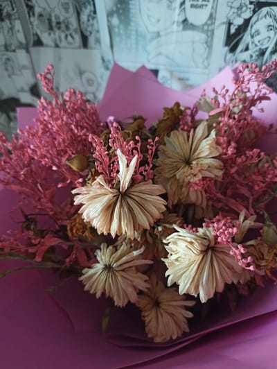 A close-up, high-angle shot shows a bouquet of dried flowers, predominantly in shades of beige and pink, arranged against a background of what appears to be manga pages. The bouquet is wrapped in vibrant pink paper. Several dried flowers with delicate, layered petals are visible, interspersed with sprigs of small pink flowers and seed pods. The manga pages behind the bouquet feature black and white drawings and text. The overall lighting is soft, creating a gentle illumination on the floral arrangement.