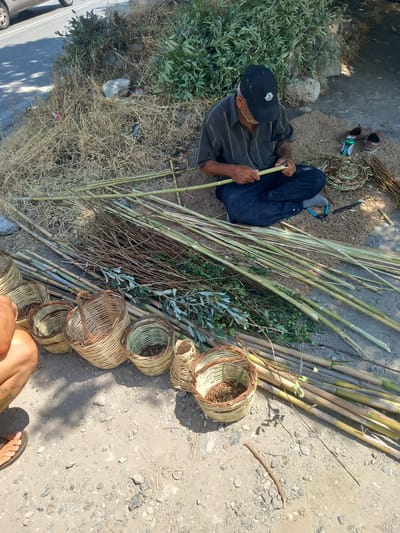 The image depicts an outdoor scene in Platani, Greece, where an elderly man is engaged in the craft of basket weaving. He is seated on the ground, wearing a black baseball cap, a striped shirt, and dark pants. In his hands, he is working with long, green stalks, presumably for basket construction. Surrounding him are numerous completed baskets of various sizes, as well as a large pile of similar stalks and dried vegetation, suggesting a crafting workshop or market stall. The ground is a mix of dirt and gravel. To the left, a portion of a car and a road can be seen, indicating proximity to a public thoroughfare. The environment is sunlit, with shadows cast by foliage, indicating an outdoor setting during daylight hours.