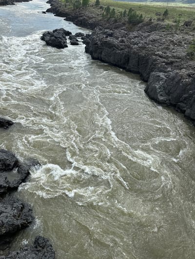 The image depicts a river with turbulent, foamy water flowing between rocky banks. The rocks are dark and jagged. On the left side of the image, some sparse vegetation is visible on a grassy area above the rocks. The water appears murky. Without additional information, it is not possible to confirm the exact location in Edigan, Russia.