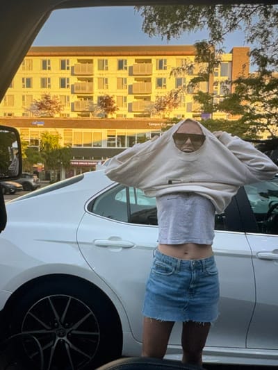 An outdoor scene is depicted from inside a vehicle, showing a young adult female interacting with her clothing next to a white sedan. The individual, seen from mid-thigh up, is extracting her head through the neck opening of an oversized, light-colored crewneck sweatshirt, which she is either donning or doffing. She wears dark sunglasses, a light grey cropped t-shirt exposing her midriff, and a light blue denim mini-skirt. Her mouth is open in an expressive manner. The white sedan's rear passenger door and wheel are visible to her left. In the background, a multi-story building with a light yellow facade and multiple balconies is prominently featured, bathed in golden natural light. Its ground level houses commercial establishments, including "Campus Drug Mart" and "Campus Dr..." signage. Trees with green and reddish foliage are interspersed throughout the scene. The location is identified as 6033 University Boulevard, Vancouver, British Columbia, Canada, which falls within Electoral Area A.