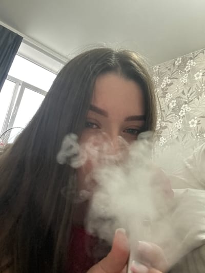 An indoor, low-angle selfie captures a young woman exhaling a large cloud of white smoke or vapor, likely from a vaping device partially visible in her right hand. Her long dark brown hair frames her face, with her eyes and eyebrows visible above the dense smoke. She appears to be looking directly at the camera. Her right hand shows manicured fingernails with light-colored polish.

The background suggests a domestic setting. In the upper portion of the frame, there is patterned wallpaper with a grey or beige floral design featuring white flowers and leaves. Below this, behind the smoke, white bedding or pillows are visible. To the lower left, a window with white frames and a dark curtain allows natural light into the room, suggesting daytime. A portion of a black fan is also visible in the lower central background. The overall impression is a casual, personal moment indoors, likely in a bedroom. No text is visible in the image.