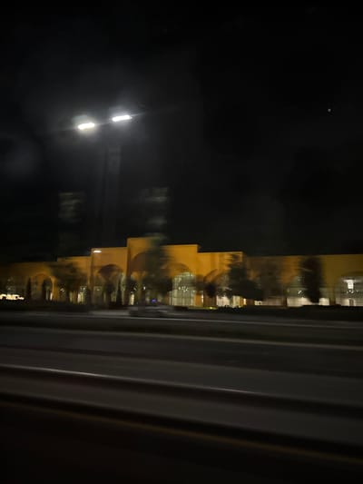 A night-time, blurred photograph captures a long, low-rise building with repeating arched windows, illuminated by interior lights. The building's facade is a warm yellow hue. Trees are visible in front of the building, appearing as dark, indistinct shapes. In the foreground, blurred horizontal lines suggest a road or highway, indicating the photo was taken from a moving vehicle. Two bright streetlights are visible in the upper left of the image, their light also appearing blurred. The overall environment is dark and urban. The building is located in Tukaytepa, Uzbekistan.