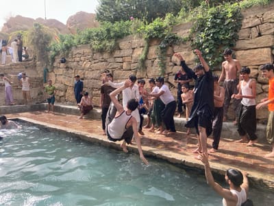 The image depicts a group of approximately 20-25 males, ranging from adolescents to young adults, engaged in recreational activity around a rectangular, light blue-green pool of water in Killi Malik Sher Mohammad, Pakistan. The scene is outdoors, under natural light, with a terraced stone wall and arid, rocky hills in the background, partially covered by green vegetation.
In the foreground, a young male in a white tank top is captured mid-jump, feet leaving the wet concrete edge, descending into the pool. Below him, another male, also in a white tank top, is partially submerged in the water, extending an arm upwards. To the right of the jumper, a male in a wet black shalwar kameez stands on the edge, arms raised high, appearing to celebrate or cheer. Numerous other males stand along the pool's edge; some are bare-chested or in wet clothing (tank tops, t-shirts, shalwar kameez), observing the activity with expressions ranging from smiling to focused attention. One individual sits on the edge, looking down at a small object in his hands. Further back and to the left, additional individuals are visible on higher stone terraces, some dressed in dry clothes, observing the scene or waiting. The overall impression is one of communal leisure and interaction around the water.