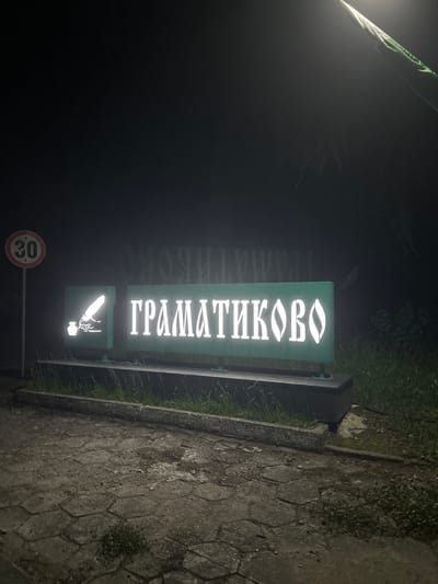 The image depicts a nighttime scene at the entrance to Gramatikovo, Bulgaria. A prominent, horizontally oriented road sign is centered in the midground. The sign consists of a dark base supporting two illuminated green panels. The right and larger panel displays the Cyrillic word "ГРАМАТИКОВО" in stylized, glowing white letters. To its left, a smaller square panel features a glowing white emblem of a quill pen standing in an inkwell.

To the left of the main sign, a red-bordered circular speed limit sign indicates "30," mounted on a slender pole. The ground in the foreground consists of wet, irregularly shaped paving stones, suggesting recent precipitation, with scattered patches of small green vegetation. A strip of unkempt grass runs along the base of the sign structure and extends to the right. The background is largely dark, with faint outlines of trees or bushes visible through the limited ambient light. A bright, upward-angled light source is visible in the upper right, illuminating a partially seen green and white flag or banner. No individuals or active subjects are present.