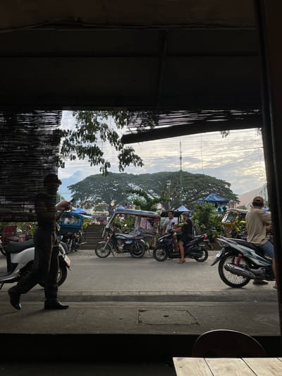 A wide shot captures a street scene in the city of Valencia, Philippines, viewed through a dark, slatted window or screen. The foreground is dominated by the dark, silhouetted interior of a building, with the slatted screen obscuring the view.

In the mid-ground, a street is lined with several motorcycles and motorized tricycles. A person in dark clothing is walking on the sidewalk on the left, looking at their phone. Two people are seated on a blue motorcycle, and another person is on a blue and black motorcycle to their right. Further down the street, a yellow and blue tricycle is parked.

The background reveals a lush green tree canopy with a mountain or large hill behind it. The sky is cloudy, with visible power lines and a communication tower. A few small blue tents or awnings are visible among the trees, suggesting a market or event. The overall lighting suggests it is either early morning or late afternoon.