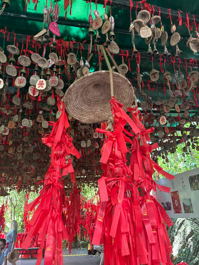 This image depicts a scene at the Sandao, China location, likely a temple or spiritual site. The foreground is dominated by hanging bundles of bright red ribbons tied with bows, with some ribbons featuring gold lettering. Behind these ribbons, the ceiling is covered with numerous small, circular wooden or metal charms and bells suspended by red strings. Some charms have symbols or writing on them, and a few pink, oval-shaped objects are also visible. A woven straw disk hangs in the mid-ground. In the background, lush green foliage is visible through gaps in the hanging objects, suggesting an outdoor or semi-outdoor environment. A poster with images and text is affixed to a rocky surface on the right side of the image. The scene conveys a sense of ritual or devotion, with the abundance of charms and ribbons suggesting wishes or prayers.