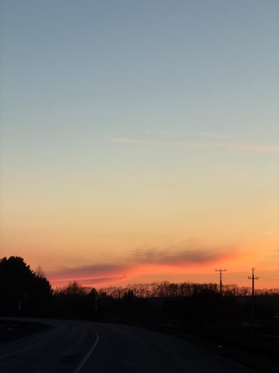 The image shows a sunset over a road near Gory, Russia. The sky transitions from light blue to orange, with a distinct pinkish band near the horizon. Silhouetted trees and power lines are visible on the right. The road curves into the foreground, disappearing into shadow.