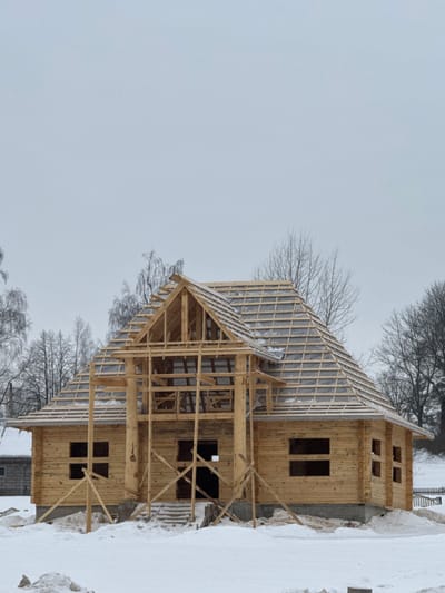 The image depicts a two-story wooden house under construction in Крэўскі сельскі Савет, Belarus, under an overcast sky during winter. The ground is covered in white snow. The house is constructed from light-colored logs, with an exposed gray concrete foundation. Multiple rectangular openings serve as provisional windows and a main entrance, lacking frames or glass. A complex, multi-gabled roof structure is evident, featuring exposed wooden rafters and a partial protective underlayment, lightly dusted with snow. A prominent A-frame gable with visible wooden framing stands over the front entrance area. Temporary wooden support beams are positioned around the front porch structure. In the background, bare deciduous trees are visible on both sides of the house, and a small, low-lying snow-covered wooden structure is partially visible to the far left. A snow-covered fence line and a gentle upward slope of snow are visible to the right. No individuals are present.