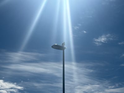The image shows a lamppost against a bright blue sky with wispy white clouds. Sunbeams are visible, creating a lens flare effect. A single bird is perched on top of the lamppost. The setting appears to be outdoors, and given the information that the location is Sund, Norway, it is likely an urban or semi-urban area. There are no people or any other significant objects or actions visible in the frame.