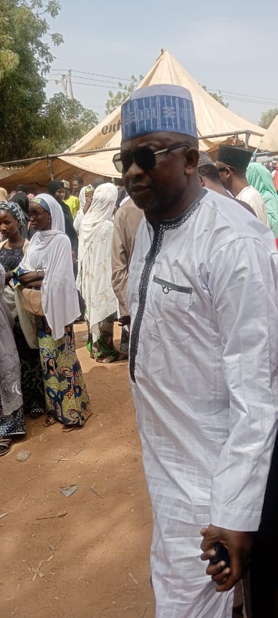 The image captures an outdoor scene under clear daylight in Sani Mai Nagge, Nigeria, featuring a gathering of people. In the immediate foreground, a dark-complexioned male subject is positioned off-center to the right, wearing a white long-sleeved tunic with intricate black embroidery on the placket and collar, dark sunglasses, and a blue-patterned kufi cap. His right hand is visible, gripping a dark-colored mobile phone.
Behind the primary subject and extending into the midground, a crowd of individuals is present, mostly women, many adorned in traditional attire including white hijabs and colorful patterned dresses. One woman in the midground wears a white hijab, glasses, and a brightly patterned dress featuring blue, yellow, and green motifs. The ground surface is unpaved, composed of reddish-brown soil with scattered small sticks and debris. In the background, a large, light-colored canvas tent with a visible "ent..." logo and partial text "WANO ST." provides shade. Overhead, utility poles and power lines are discernible against a pale blue sky, with sparse green trees flanking the scene. No specific actions or direct interactions are clearly depicted beyond individuals standing within the gathering.