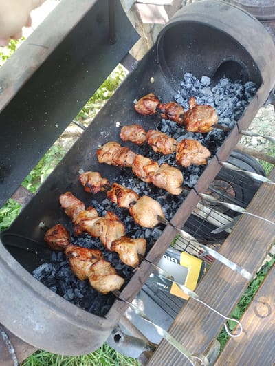 In Kniazhdvir, Ukraine, a charcoal grill is filled with glowing embers. On metal skewers, pieces of meat are being grilled to a golden brown color. The grill itself is made from a large metal barrel, tilted on its side, with a wooden plank placed underneath to support it. Several skewers are already loaded with meat, and some are arranged in rows over the coals. The surrounding area appears to be outdoors, with green grass and some greenery visible in the background. A hand is partially visible at the top of the frame, suggesting someone is tending to the grill.