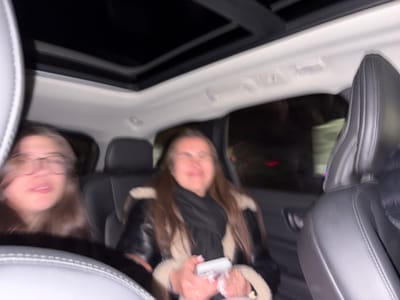 Inside a car at night, two women sit in the back seat. The woman on the left wears glasses and has long hair. The woman on the right has long brown hair and wears a black jacket with a light-colored fuzzy trim and a black scarf. She holds a white object in her hands. The car's interior includes black leather seats and a sunroof. The image is somewhat blurry, suggesting motion. A dark exterior can be seen through the window.