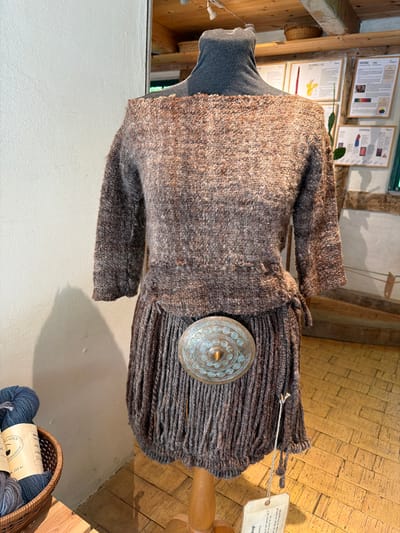 This image shows a display of knitted garments, likely in a shop or workshop, located in Lejre, Denmark. The central focus is a mannequin wearing a brown, textured knitted top with a wide neckline and three-quarter length sleeves. The top is cinched at the waist with a tie on the right side. Below the top, a separate knitted skirt with a fringed hem is displayed. A large, ornate circular metal buckle with intricate patterns in shades of blue and bronze is fastened at the waist over the skirt. To the left of the mannequin, a woven basket contains several balls of dark blue and gray yarn. In the background, shelves are visible with various items, including what appear to be woven baskets and framed informational posters related to crafts. The environment appears rustic, with wooden beams on the ceiling and wooden floorboards. The lighting suggests an indoor setting.