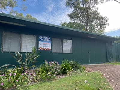 An outdoor, eye-level, landscape-oriented photograph depicts a single-story, dark green building, likely a community hall or hut, set in Riverhills, Australia. The building is constructed with a corrugated metal section on the far left and flat panels making up the rest of its facade. Several large, rectangular windows, covered with a protective mesh or grille, are visible along the front.

In front of the building, a well-maintained garden bed features various green plants and flowering shrubs with small pink blossoms. Beyond the garden, a grassy area with some scattered leaves extends to the left, where a paved path with interlocking bricks is partially visible. The scene is illuminated by bright daylight under a clear blue sky with some scattered white clouds. A large leafy tree, possibly in the foreground or reflected, is prominent against the sky on the left side of the image.

A blue and white sign is affixed to the right side of the building, next to a window. The text on the sign, oriented vertically, reads:
"Middle Park Girl Guides"
"HUT FOR HIRE"
"THE GIRL GUIDES"
"www.girlguidesqld.org"
"mail@girlguidesqld.org"
The Girl Guides trefoil logo is also visible on the sign. Below this, in smaller text, it states "© 2013 The Guide Association. Registered Charity No. 306016." The sign indicates the building serves as a Girl Guides hut and is available for hire. No people or activities are visible.