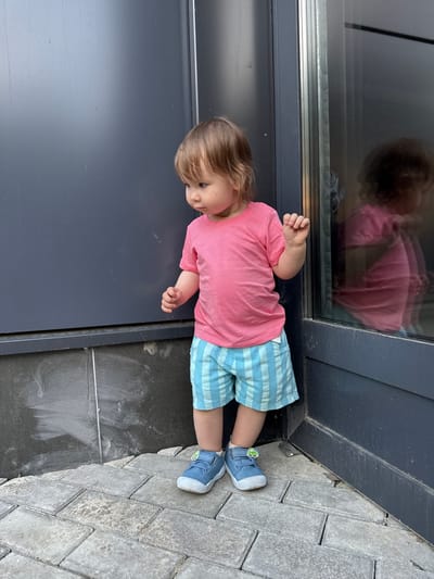 A young child stands near the corner of a building with dark gray siding and a large glass window, possibly in the city of Kraskovo, Russia. The child is wearing a bright pink t-shirt and blue and white striped shorts, with blue sneakers adorned with small green car charms. The child's gaze is directed downward and to the left. The ground is paved with gray interlocking bricks. A reflection of the child in the glass window shows them wearing the same clothing. The scene suggests an outdoor urban environment, with the building's architecture indicating a modern construction.