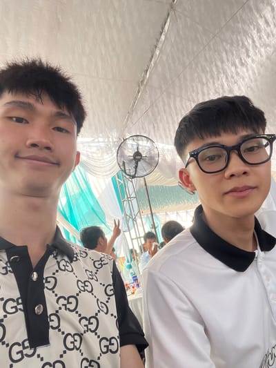 This image is a selfie featuring two young men at what appears to be a social gathering or event in Bình An, Vietnam. The photo is taken from a low angle, looking upwards.

In the foreground, the top subject is a young man with short dark hair, wearing black-rimmed glasses and a white polo shirt with black trim. He looks directly at the camera with a neutral expression. Below him, the second young man, also with short dark hair, smiles slightly while looking into the camera. He wears a white short-sleeved shirt patterned with a black interlocking 'G' logo.

The background reveals an event space adorned with white and light blue/teal fabric drapes. A black industrial-style fan is visible, hanging in the mid-ground on the left. The ceiling is covered with white pleated fabric, suggesting a temporary structure or marquee. Several other individuals are present in the background, out of focus; one person on the right is seen making a peace sign. On a table further back, small packaged items, possibly food or drinks, are visible. The bright lighting suggests it is daytime. The presence of the fan implies warm weather conditions. The overall atmosphere appears casual and friendly.