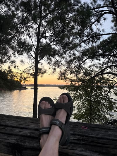 A low-angle, medium shot captures a serene sunset scene at Lake Lanier, located in Sugar Hill, Georgia, United States. The foreground is dominated by a pair of feet wearing dark, strapped sandals resting on a weathered wooden surface. The feet are positioned to the left and center of the frame, with the left foot angled slightly towards the viewer. Behind the feet, the tranquil waters of the lake stretch out, reflecting the warm hues of the setting sun, which casts a gradient of orange, yellow, and blue across the sky. The horizon is dotted with the silhouettes of trees lining the distant shore. Pine trees, with their characteristic needles and branches, frame the upper portion of the image, creating a dappled canopy against the colorful sky. The overall atmosphere is one of peace and relaxation, with nature's beauty taking center stage.