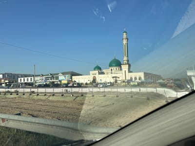 A wide-angle, eye-level shot captures a modern mosque in Baxt, Uzbekistan. The mosque features a tall, slender minaret with green and white stripes, topped with a green dome. Two smaller green domes are visible on the main building. The mosque's facade is white with decorative arches. In front of the mosque, a paved area is filled with several parked cars, including white vans and dark sedans. Beyond the mosque, a line of multi-story buildings with flat roofs stretches across the background. The scene is framed by a reflective surface, likely a car window, which casts streaks of light and reflections across the image. In the foreground, a muddy construction site with visible tire tracks is separated from the mosque area by a black metal fence. The sky is clear and blue, indicating fair weather.