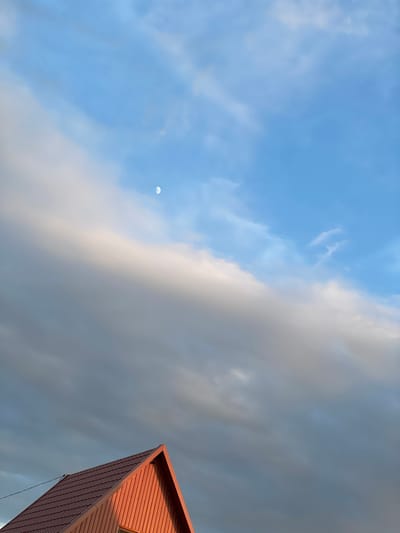 The image captures a dramatic sky scene with a crescent moon visible in the upper left quadrant. The sky is divided between a bright, clear blue on the left, dotted with wispy white clouds, and a darker, more overcast expanse of grey clouds on the right. The division creates a striking contrast. In the lower right corner of the image, the sharp, angular edge of a modern building with reddish-brown siding is visible, reaching towards the sky. The building's architecture is geometric and contemporary. The lighting suggests either early morning or late afternoon, as the sun casts a warm glow on the side of the building, while the sky exhibits a mix of daylight and approaching or receding clouds. There is no visible text in the image. The overall impression is one of a vast, dynamic sky meeting the man-made structure, creating a sense of scale and perhaps a hint of atmospheric grandeur. The weather appears to be changing, with a mix of sunshine and cloud cover. The location is identified as Buynaksk, Russia.