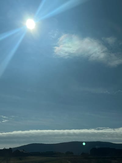 The image displays an outdoor landscape scene under a bright, predominantly light blue sky, identified as being in the Town of Fishkill, United States of America. The sun, a bright white orb, is prominently positioned in the upper left quadrant, emitting strong, diagonal lens flares across the upper left portion of the frame. Scattered, wispy white clouds are visible in the upper right quadrant, with some iridescent coloration at their edges. A small, dark, indistinct object, possibly an avian subject or insect, is observable against the clear blue sky in the upper-middle section. Along the lower horizon, a dark, undulating silhouette of a mountain range is visible, partially obscured by a distinct, horizontally elongated band of white and gray clouds immediately above it. Below the mountain range, in the immediate foreground, are dark, indistinct silhouettes of multiple buildings and open ground, indicating structures such as houses or barns within an open field environment. A prominent circular green optical artifact is visible against the dark mountain silhouette in the lower right quadrant, approximately centered horizontally. No human subjects or discernible actions or interactions are present in the scene.