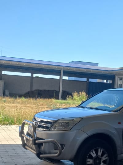 The media file shows a close-up view of the front right side of a silver SUV parked on a paved area. The vehicle has a chrome bull bar attached to the front bumper and a visible tire. Behind the vehicle, there is a building with a blue corrugated metal roof supported by white beams, and an open-sided structure with concrete walls. The ground surrounding the building appears to be dry earth with some vegetation. The sky is clear and blue, suggesting it is daytime. The scene appears to be an outdoor setting, possibly a residential or commercial area in Yorkatay, Uzbekistan. No people are visible in the image. There is some text on the license plate of the vehicle, but it is not clearly legible. The overall impression is of a sunny day.