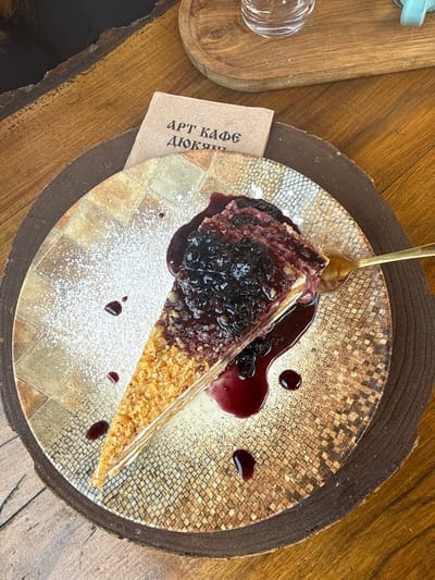 A slice of layered cake, topped with a dark berry sauce and fresh berries, sits on a patterned plate on a wooden table. A gold fork is inserted into the cake. A small brown card with Cyrillic text is placed behind the plate, and a glass is visible in the background on another wooden surface. The scene is located in the city of Shiroka Laka, Bulgaria.