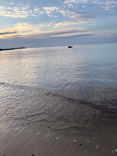 The image shows a serene seascape at Rohuneeme, Estonia, under a partly cloudy sky. The foreground features a sandy beach with small waves lapping at the shore, creating ripples and foam. In the distance, the calm sea stretches towards the horizon, reflecting the soft hues of the sky, which are painted with pastel shades of blue, pink, and yellow. A few scattered rocks are visible in the water, and a distant ship can be seen on the horizon. The overall atmosphere is tranquil and peaceful, capturing the beauty of a coastal sunset.
