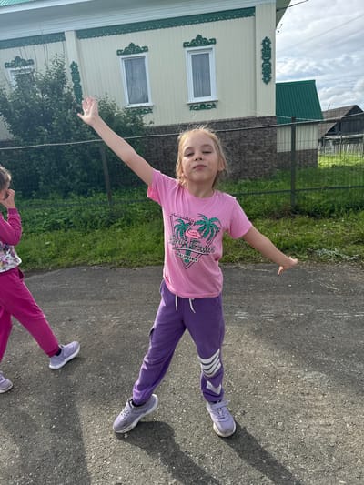 In Bolshoy Turysh, Russia, a young girl with blonde hair and light skin is standing outdoors on a paved surface, facing the camera. She is wearing a pink t-shirt with a graphic design and purple sweatpants. Her right arm is raised, with her hand open. To her left, another child, partially visible, is wearing pink sweatpants. The background shows a light-colored building with green trim, a green fence, and lush green vegetation. The sky above is partly cloudy. The overall scene appears to be a residential area on a daytime, possibly summer, day.