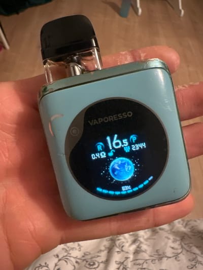 The scene, set in Montreux, Switzerland, features a light blue Vaporesso brand vaping device held in a human hand. The device exhibits visible wear, scuffs, and minor paint chips on its casing and silver-colored trim. A clear plastic pod with a black mouthpiece is inserted at the top, containing a visible amount of e-liquid. The device's circular digital display is illuminated, showing "VAPORESSO" at the top, "16.5s" (likely puff duration), "0.4Ω" (coil resistance), "2344" (puff count), a glowing blue Earth graphic in the center, and "83%" battery life indicated by a percentage and an arc of blue segments. The hand holding the device shows skin texture, with fingers curled around its sides and the palm partially visible beneath. The background is blurred, revealing a patterned fabric, possibly a bedsheet, with light grey floral or damask motifs in the lower right portion of the frame, and an indistinct lighter surface further in the background.