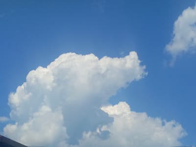 The image shows a bright blue sky filled with fluffy white cumulus clouds. The clouds are voluminous and appear to be in motion. In the lower left corner, the dark silhouette of a sloped roof or structure is visible, partially obscuring the sky. The scene is outdoors and depicts a clear day. The image is a photograph of the sky above Novomikhaylovsky, Russia.