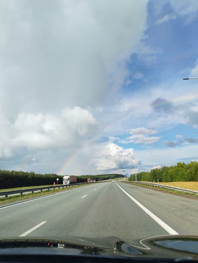 The image captures a view from inside a vehicle driving on a highway in Russkie Kirmeni, Russia. The sky is a mix of dramatic, dark storm clouds and bright blue patches, with a visible rainbow arc across the sky. Trees line both sides of the highway, with a grassy field bordering the right side. Several trucks are visible on the highway, with one white truck prominently featured in the foreground. The road is paved asphalt with white lane markings. The lower portion of the image shows the dashboard and hood of the vehicle.