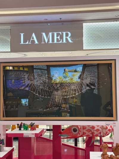 The image shows a storefront display featuring the brand "LA MER". Above the display, the "LA MER" logo is prominently displayed in large, white, sans-serif font against a dark background. To the left and right of the logo are patterned screens. Below the logo, a large framed display showcases a digital advertisement with an artistic depiction of a bird's wings and a hand holding a globe. The advertisement is reflected in the glass, showing the interior of a store with shelves and products. In the foreground, there are various decorative items on pink pedestals. These include small figurines resembling animals and a colorful, elongated fish-shaped kite or decoration with red and white patterns. A sign with Chinese characters is visible above the fish decoration. The overall environment appears to be an indoor shopping area, likely a mall, given the lighting and display setup. Based on the visible Chinese text on the signs and the typical branding of stores in such locations, it is plausible this scene is located in Jianye District, China.
