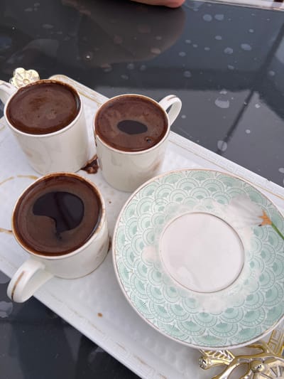 In an overhead, close-up shot, three cups of dark, frothy coffee sit on a white, rectangular tray with gold trim. The tray is placed on a glossy black surface, possibly a table, which has a speckled, wet appearance. To the right of the cups is a decorative saucer with a pale green, fan-like pattern and gold accents, also featuring a white floral design. One cup has a gold flower-shaped lid. The setting appears to be outdoors, possibly on a patio or balcony, given the bright lighting. There are no individuals visible in the frame, and no specific location in Novy, Russia can be identified.