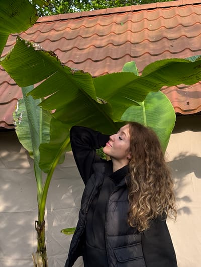 A woman with curly brown hair is resting her head on a large green banana leaf. She is wearing a black jacket. The banana leaf is next to a roof covered in red tiles. The background is a beige fabric. The location is not discernible from the image.