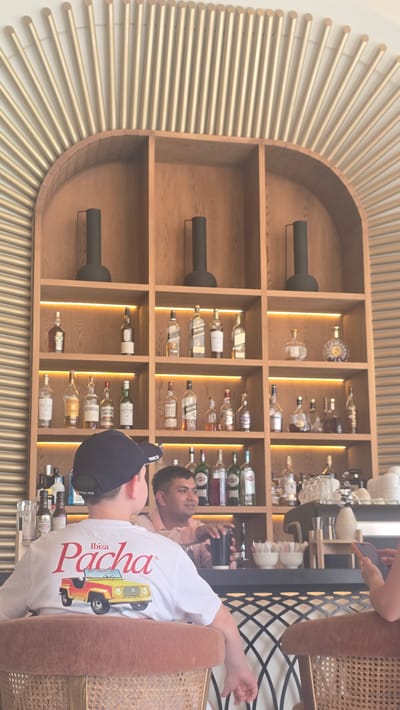 This media file shows a bar or restaurant setting with a large, ornate wooden shelf filled with bottles of alcohol. A man wearing a white t-shirt with the text "Pacha Ibiza 1992" and an image of a yellow jeep is sitting in a chair, leaning back and looking away from the camera. He is wearing a black baseball cap. Another person, partially visible in the background, is looking at their phone. The shelf is illuminated by warm LED lights, and the bottles on display appear to be various spirits and liqueurs. The overall aesthetic suggests a stylish and well-stocked establishment, possibly in a Mediterranean location given the "Ibiza" text. The lighting suggests it could be evening or indoors with artificial lighting.