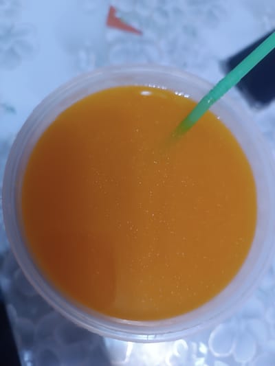 A top-down close-up shot shows a clear plastic cup filled with a vibrant orange liquid, likely juice. A green plastic straw with segmented ridges is partially submerged in the liquid, extending from the upper right towards the center. The surface of the liquid has tiny white specks scattered across it, possibly pulp or air bubbles. The cup's rim is visible along the bottom and left edges of the frame. The background is a blurred, light blue surface with a subtle floral pattern, suggesting a table or placemat. The lighting creates subtle reflections on the liquid and the plastic cup. No people or specific urban features of Бидайкөл, Kazakhstan are discernible in this image.
