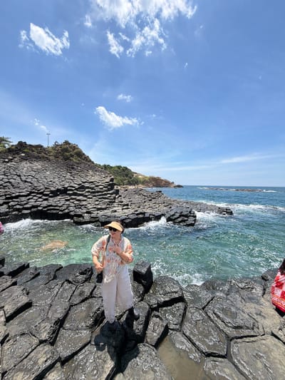 In a sun-drenched outdoor scene, a person stands on a unique geological formation of dark, hexagonal basalt columns near the ocean. The sky above is a clear, bright blue with scattered white clouds. The turquoise water laps against the rocks, creating small waves and white foam.
The person, facing away from the viewer and looking towards the water, wears a light-colored floral shirt and white pants, accessorized with a straw hat and sunglasses. They are holding a small purse with a chain strap. The hexagonal basalt columns, a notable geological feature, are wet and shiny, reflecting the sunlight.
The overall atmosphere suggests a pleasant day for sightseeing and enjoying the natural scenery. The location appears to be a coastal area with volcanic rock formations. The time of day is likely midday due to the bright sunlight and clear sky. No visible text is present in the image.