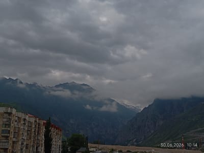 The image depicts a cloudy sky over a valley with mountains. In the foreground, a multi-story apartment building is visible on the left. The mountains in the background are covered in greenery, with some snow-capped peaks. A date and time stamp, "30.06.2026 10:14", is present in the lower right corner. The scene appears to be of the city of Tyrnyauz, Russia. There are no people or animals visible in the image. The environment is natural and urban, with the apartment building contrasting with the mountainous landscape.