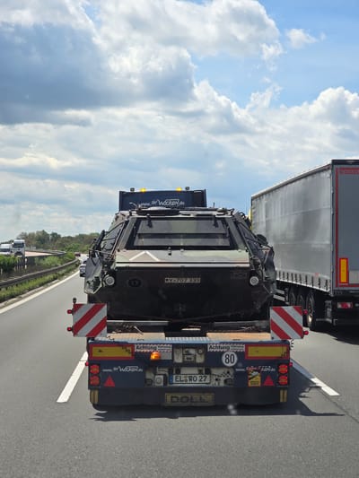 The image shows the rear view of a flatbed truck carrying a military-style armored vehicle on a highway. The truck is in the right lane, and a large semi-trailer truck is to its right. The sky is partly cloudy. The armored vehicle is dark green with a camouflage pattern and has the license plate "Y-707 339". There are several warning labels and lights on the back of the flatbed trailer, including one that says "Achtung Fahrzeug schert aus" (Caution vehicle swerves) and a speed limit sign for 80. Another sign says "ATTENT ON ANGLES MORTS" which translates to "Attention Blind Spots". The website "www.erwocken.de" is visible on the trailer. The highway appears to be a German autobahn, based on the signage and the presence of a KOGEL trailer. The specific location of Dessau, Germany, is not explicitly indicated in the image, but the overall context suggests it is a German highway. There are no people clearly visible in the image. The scene depicts transportation of a heavy vehicle on a highway.