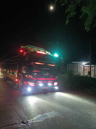 This nighttime photograph depicts a large, heavily customized truck driving on a paved road. The truck, likely a flatbed or a general cargo vehicle with high side walls, is the dominant subject, captured with motion blur that emphasizes its movement. It features numerous bright lights: powerful white headlights, red marker lights along its cabin and cargo area, and green lights on its roof, creating vibrant light trails and reflections on the wet or textured asphalt.
The scene is set under a dark, clear night sky, with a prominent, bright full moon visible in the upper left. To the right of the road, the dark silhouettes of trees and dense foliage are present, with some tree branches extending into the upper right corner of the frame. In the mid-ground on the right, a simple building with visible brick and corrugated material construction can be seen, partially illuminated by ambient light. The road surface in the foreground shows detailed texture, tire marks, and bright illumination from the truck's headlights, highlighting a small dark patch of liquid or debris near the bottom center. The image conveys a sense of a journey at night in a rural or semi-rural area, consistent with the Meuse region of Indonesia. No discernible text is visible on the truck or in the surroundings.