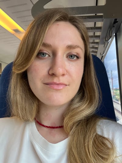 A woman with blonde, wavy hair and blue eyes is depicted in a close-up selfie, seated within a modern train interior in Stara Pazova Municipality, Serbia. She appears to be in her 20s or early 30s, with fair skin exhibiting minor irregularities, including small bumps and redness on her forehead and cheeks. Her lips are glossy, and she wears light eye makeup. She is dressed in a white crew-neck t-shirt and a red choker necklace composed of small, fragmented beads. Reddish patches are visible on her neck above the necklace. She gazes directly at the camera with a neutral expression. The background features a dark blue upholstered seat headrest to her left and a window to her right, through which train tracks, green landscape, and a distant utility pole are visible under a cloudy sky. The train's ceiling, with recessed warm yellow lighting and perforated panels, is partially visible above her.
