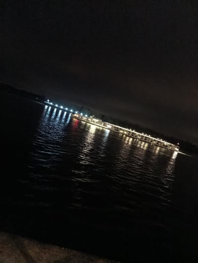 The image shows a night view of the Rhine River in Emmerich, Germany. A long, brightly lit passenger cruise ship is traversing the water. Reflections of the ship's lights are visible on the river's surface. In the distance, the far shore of the river is faintly visible, marked by a series of lights. The sky is dark and overcast. The photograph appears to have been taken from a low vantage point, possibly near the riverbank.