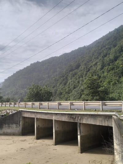 This media file captures a scenic view of a roadside underpass situated at the base of a lush, tree-covered mountain. The underpass, constructed from concrete, features multiple arched openings that lead to a sandy riverbed or dry creek bed below. The sandy terrain is marked with footprints.

Above the underpass, a paved road runs horizontally across the frame, bordered by a guardrail. A single person, appearing as a distant figure, is walking along the roadside. The road is flanked by green grass and some scattered debris to the left.

The background is dominated by a steep mountain slope densely covered with a variety of green trees, creating a thick forest canopy. A series of overhead power lines stretch across the sky, adding a man-made element to the natural landscape.

The sky is overcast with clouds, suggesting a possibly humid or approaching rainy weather. The overall lighting is diffused, indicating it could be morning or late afternoon, but the overcast conditions make it difficult to determine the exact time of day. The location is identified as Meudheun, Indonesia, which aligns with the tropical vegetation visible in the image. The scene evokes a sense of natural beauty and the coexistence of infrastructure with the environment. No visible text is present in the image.
